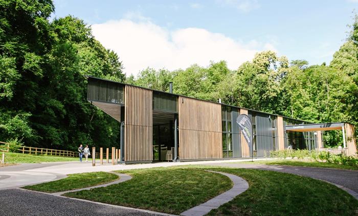 view of a modern single storey building. It has full height windows and vertical wooden cladding. in front of the building is a grassed area with path
